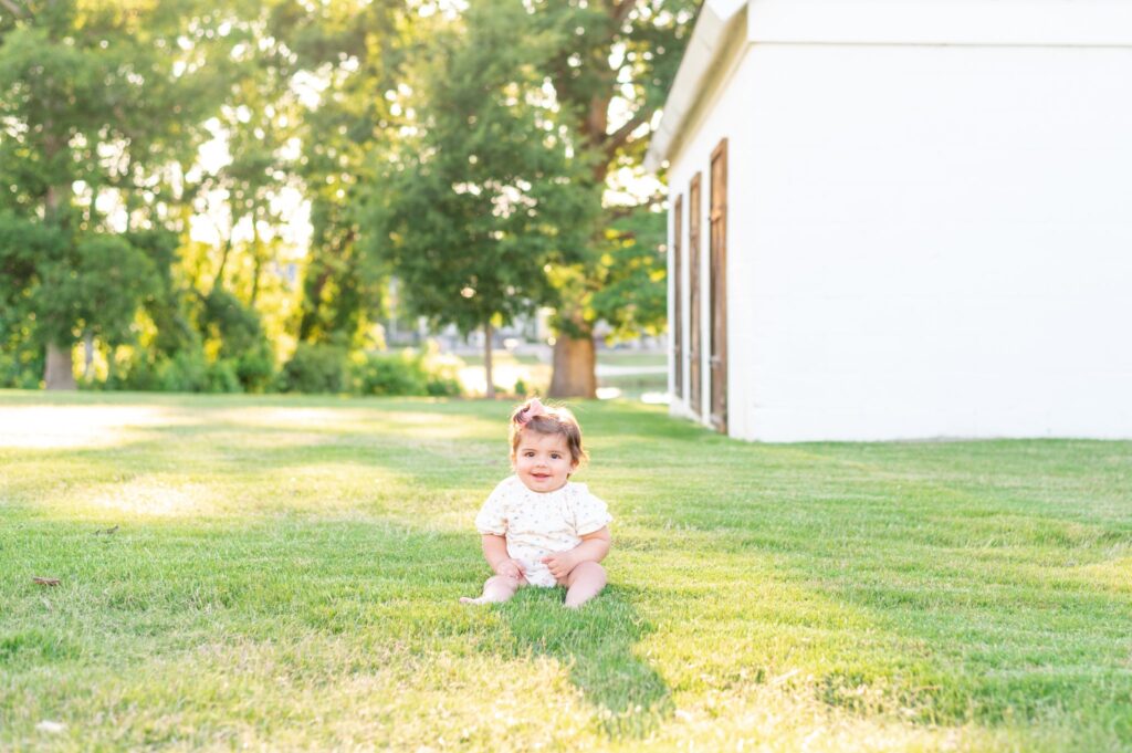 little baby happy smiling at the camera  sitting in a field with a white barn behind her